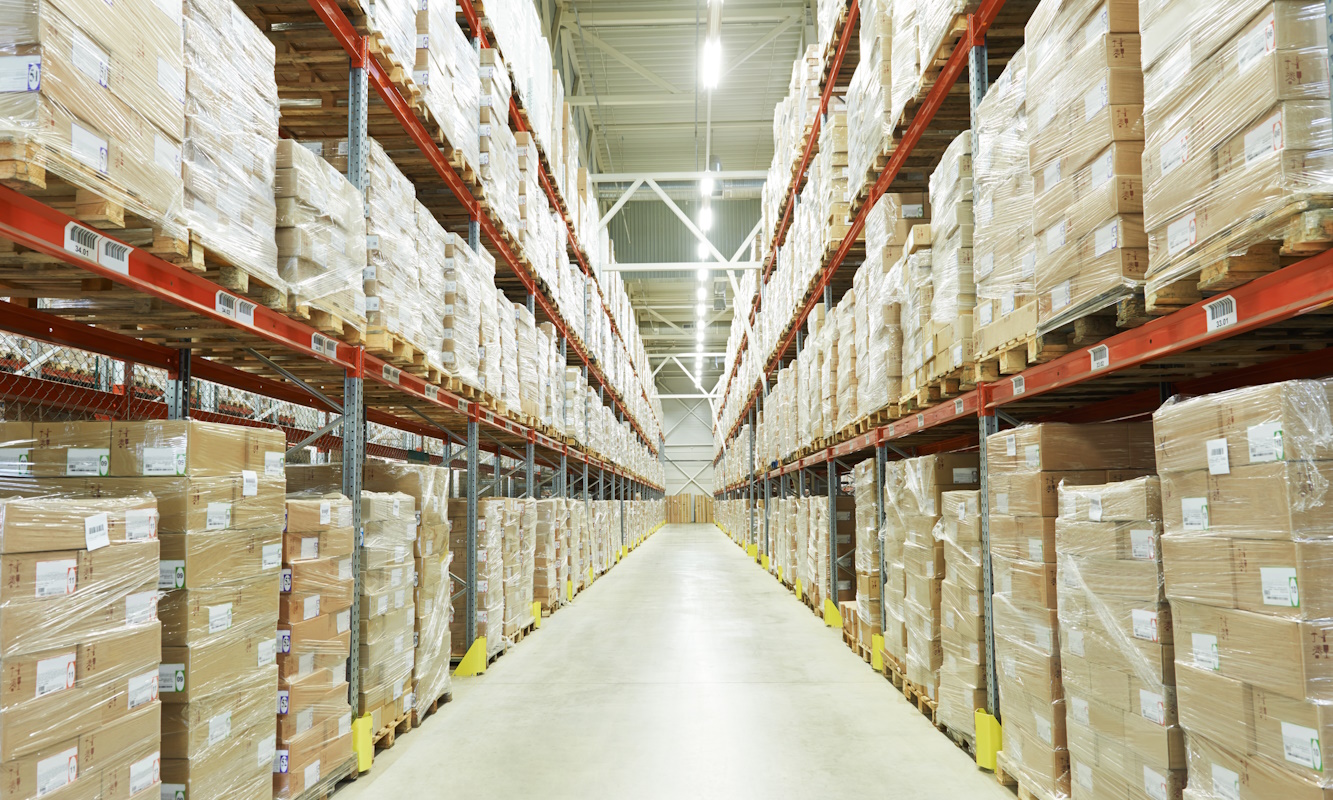 Interior view of a modern warehouse with tall racks filled with neatly stacked pallets wrapped in plastic, forming symmetrical aisles under bright industrial lighting.
